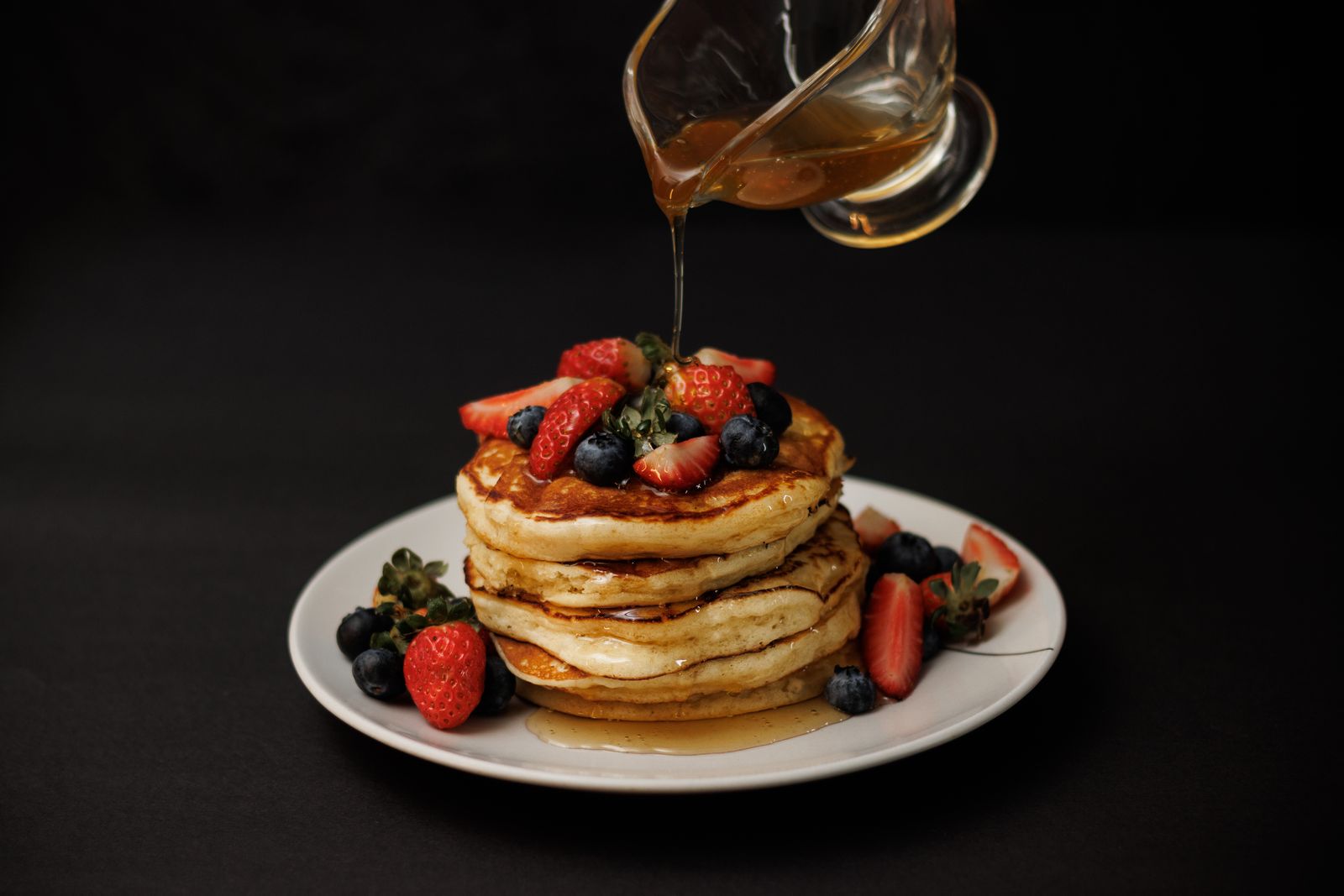 Stack of oat pancakes with berries and maple syrup being poured